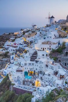 an aerial view of white buildings and windmills on the cliff above the water at dusk