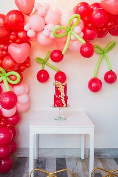 a table topped with a cake covered in red and pink balloons next to a wall