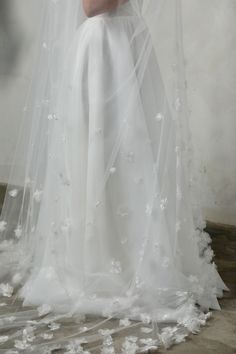 a woman in a white wedding dress and veil with flowers on the skirt is standing