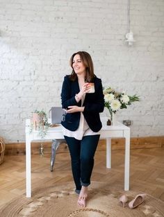 A brand photographer captures a woman standing by a white desk holding a cup, barefoot on a rug. The desk features flowers, a glass, and a phone charger. The room is adorned with a white brick wall and wooden floor. Wellbeing Coach, Brand Photoshoot