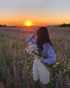 a woman standing in a field at sunset with flowers on her shoulder and the sun setting behind her