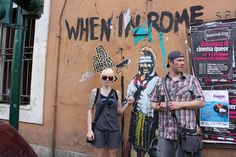 two people standing in front of a wall with graffiti on it and one person holding a camera