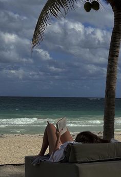 a woman laying on top of a couch under a palm tree next to the ocean