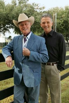 two men standing next to each other in front of a fence with trees and grass