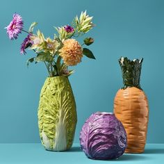 three colorful vases with flowers in them on a blue tableclothed surface, one has cabbage and the other has an onion