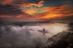 a bridge in the middle of a foggy city at sunset with clouds surrounding it