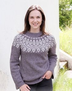 a woman standing in front of a white wall wearing a gray sweater and black pants