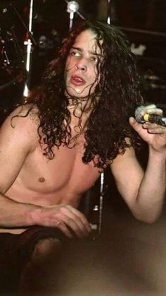 a shirtless man with long curly hair holding a cell phone in his right hand