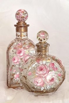 two perfume bottles with pink flowers on them sitting on a white cloth covered tablecloth