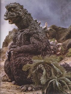 a large godzilla sitting on top of a pile of dirt next to a palm tree