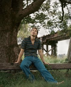 a man sitting on a wooden bench in the grass next to a tree with his eyes closed