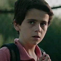 a young boy wearing a pink shirt and black backpack looking at the camera while standing in front of a fence