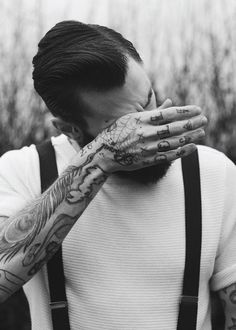 a man with tattoos covering his face and holding his hands to his mouth while standing in front of tall grass