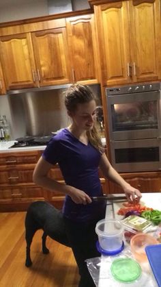 a woman cutting up vegetables on top of a kitchen counter next to a black dog