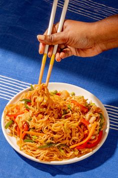 a person holding chopsticks over a plate of noodles with carrots and peppers