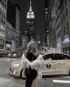 a woman standing on the side of a road in front of a car and tall buildings