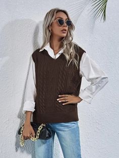 a woman standing next to a white wall wearing a brown sweater vest and blue jeans
