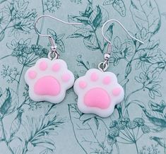 a pair of pink and white cat paw earrings on a blue background with floral designs