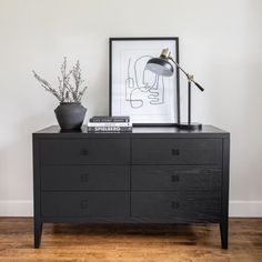 a black dresser with some books and a lamp on it next to a framed drawing