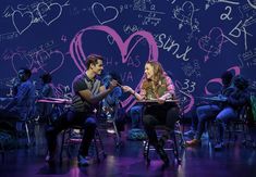 two people sitting at desks in front of a wall with writing and hearts on it