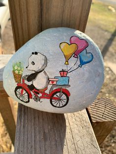 a painted rock with a panda riding a bike and balloons attached to it on a wooden fence post