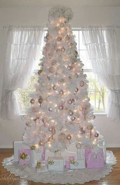 a white christmas tree with presents under it and lights on the top, in front of a window