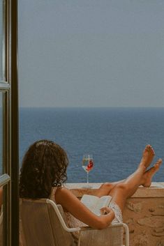 a woman sitting in a chair looking out at the ocean with a glass of wine