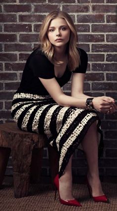 a woman is sitting on a bench in front of a brick wall and wearing red shoes