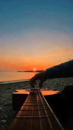 someone is playing the guitar at sunset on the water's edge with their hand