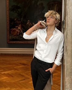 a young man leaning against a wall with his hands on his hips, wearing a white shirt and black pants