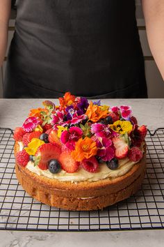 a cake with fruit and flowers on top is sitting on a cooling rack in front of a person