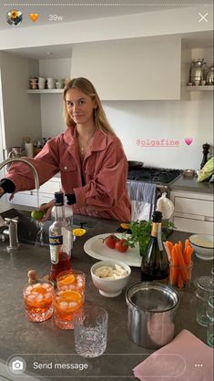 a woman standing in front of a kitchen counter filled with food and drinks, next to a bottle of wine