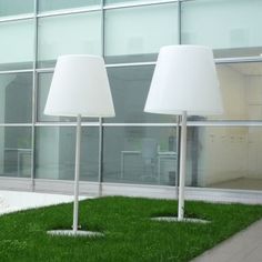 two lamps sitting on top of green grass next to a tall glass building with windows