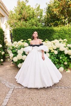 a woman in a white dress standing next to some hydranges and bushes with her hands on her hips