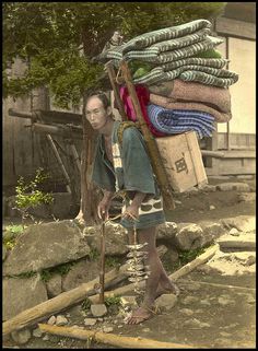 an old photo of a woman carrying luggage on her head and standing in front of a pile of clothes