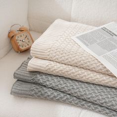 a stack of folded sweaters next to an alarm clock on a white couch with text