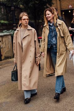 two women walking down the street in trench coats and jeans, one carrying a handbag