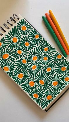 a notebook with an orange and green flower design next to colored pencils on a white surface