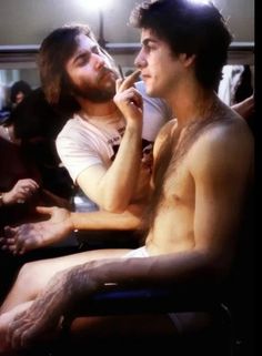 two shirtless men sitting next to each other in a room with one man drinking from a cup
