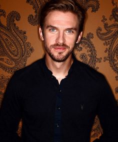 a man standing in front of a wall with a paisley pattern on it and looking at the camera