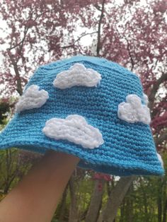 a hand is holding up a crocheted blue hat with white clouds on it