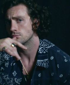 a man with curly hair wearing a blue shirt and gold ring sitting in front of a black background