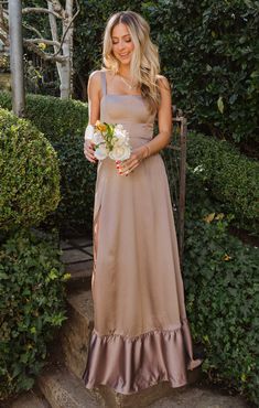 a woman standing in front of some bushes wearing a tan dress and holding a bouquet