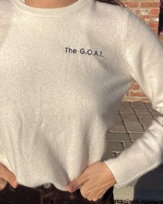 a woman standing in front of a brick wall wearing a white sweater and brown pants