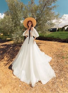 a woman in a white dress and straw hat poses for a photo on a dirt field