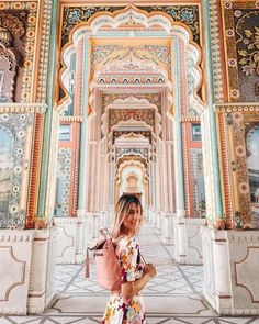 a woman standing in front of a building with colorful painted walls and arches, holding a pink backpack