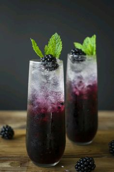 two glasses filled with blackberries and ice on top of a wooden table next to berries