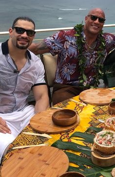 two men sitting at a table with plates of food on it, smiling for the camera