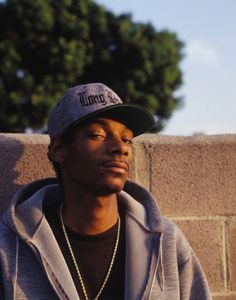 a young man wearing a hat sitting against a brick wall and looking off into the distance