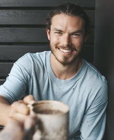 a man sitting at a table with a cup in his hand and smiling for the camera